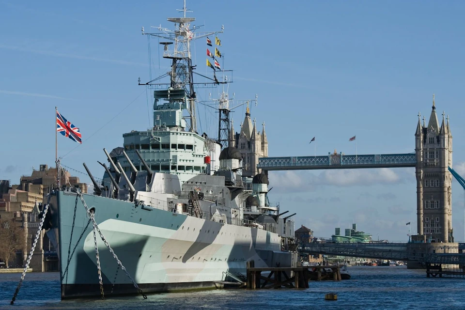 A historic battleship docked on a river with a bridge and tower in the background under a clear blue sky.
