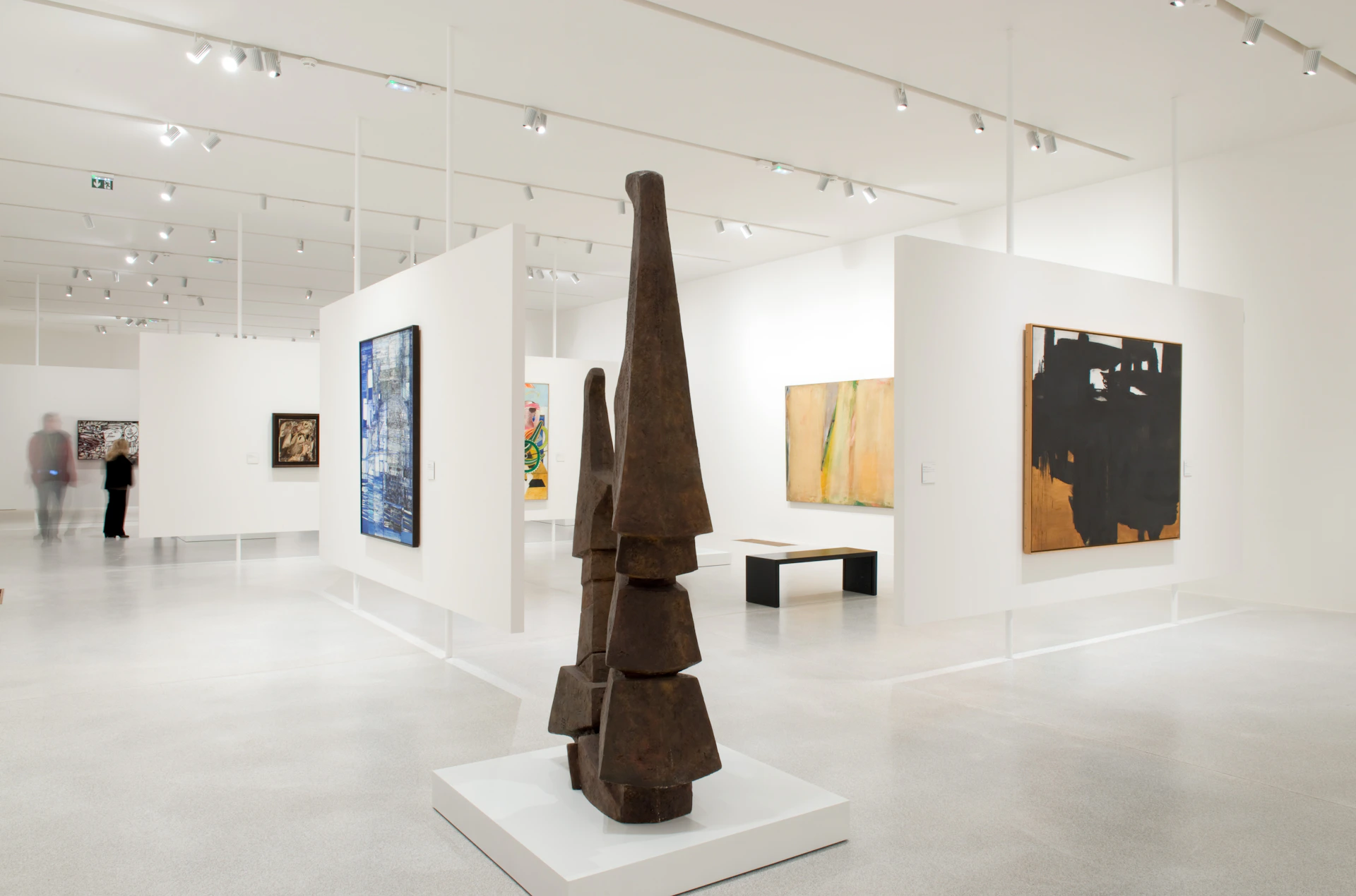 Modern art gallery interior with abstract paintings on white walls and a geometric sculpture on a pedestal in the foreground.