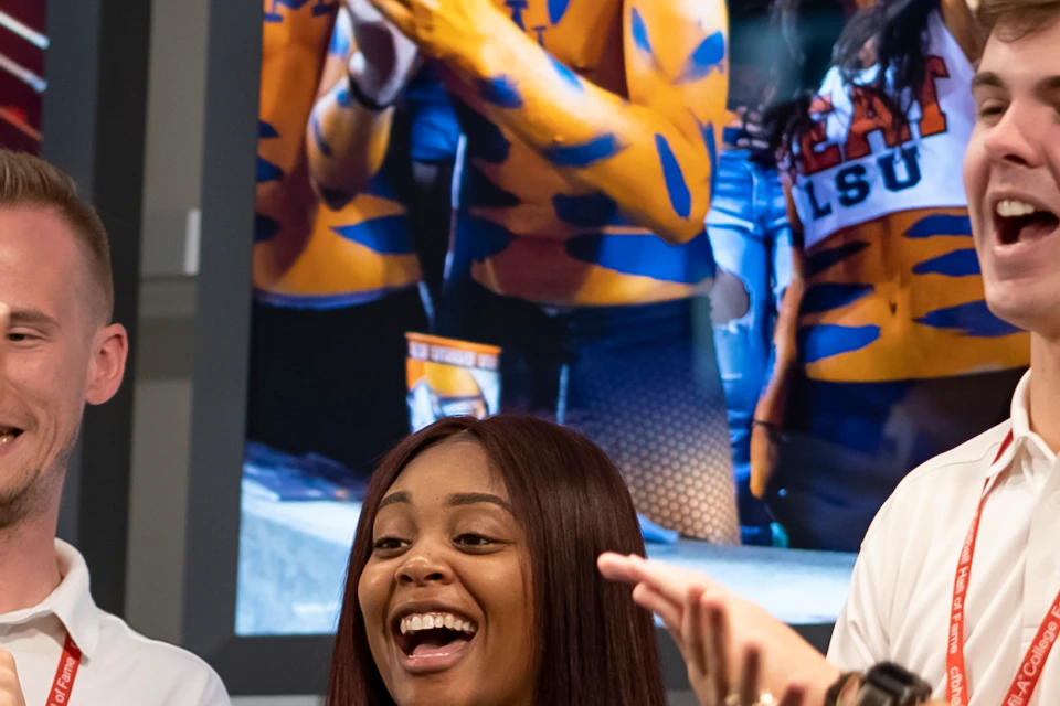 Three people with "FAN" lanyards cheer in front of a sports-themed backdrop. They appear excited and happy.