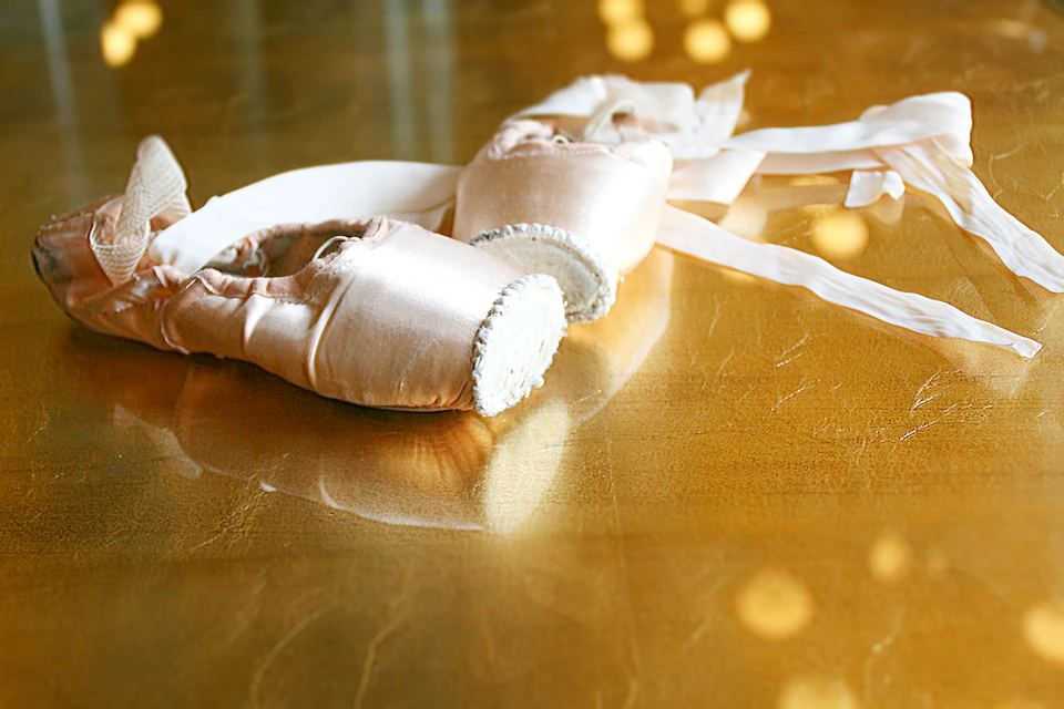 A pair of pink satin pointe ballet shoes with ribbons, placed on a reflective golden surface.