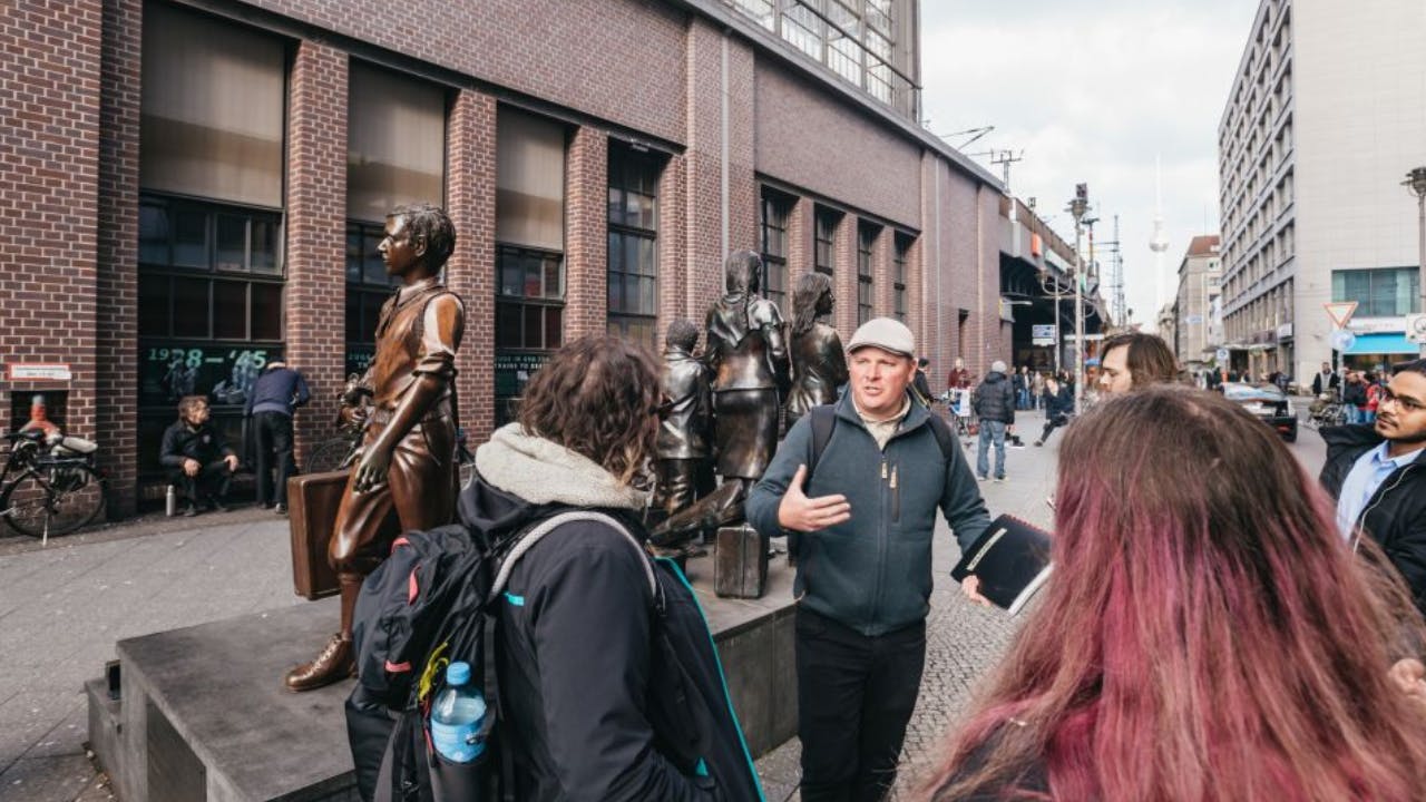A tour guide speaks to a group of people near a bronze statue on a sidewalk in a city with brick buildings and pedestrians.
