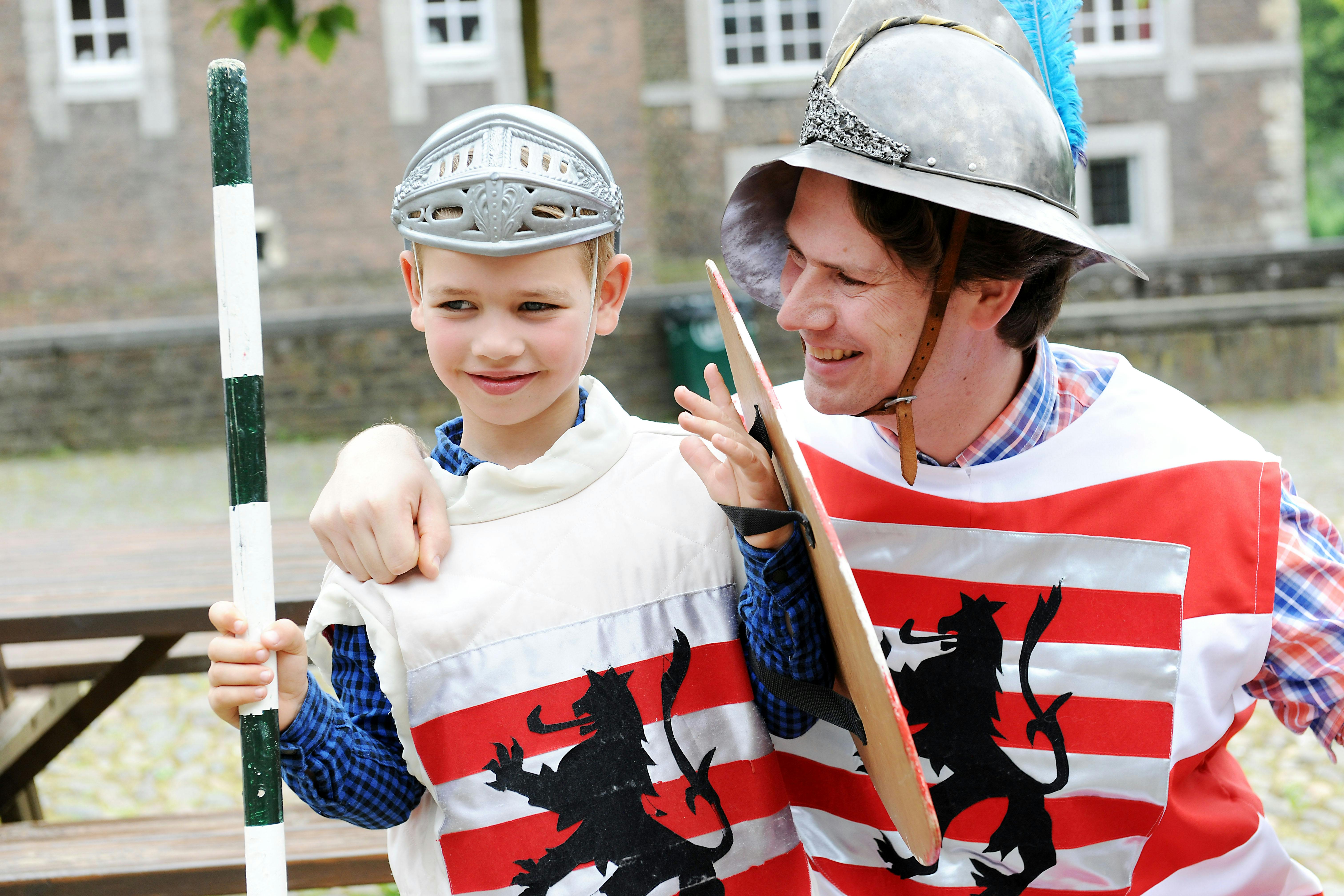A man and a boy, both wearing medieval helmets and red-striped tunics with black lion emblems, smile and stand outside.