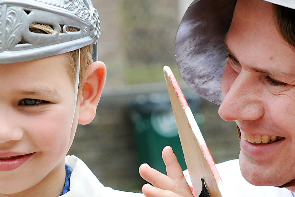 A man and a boy, both wearing medieval helmets and red-striped tunics with black lion emblems, smile and stand outside.