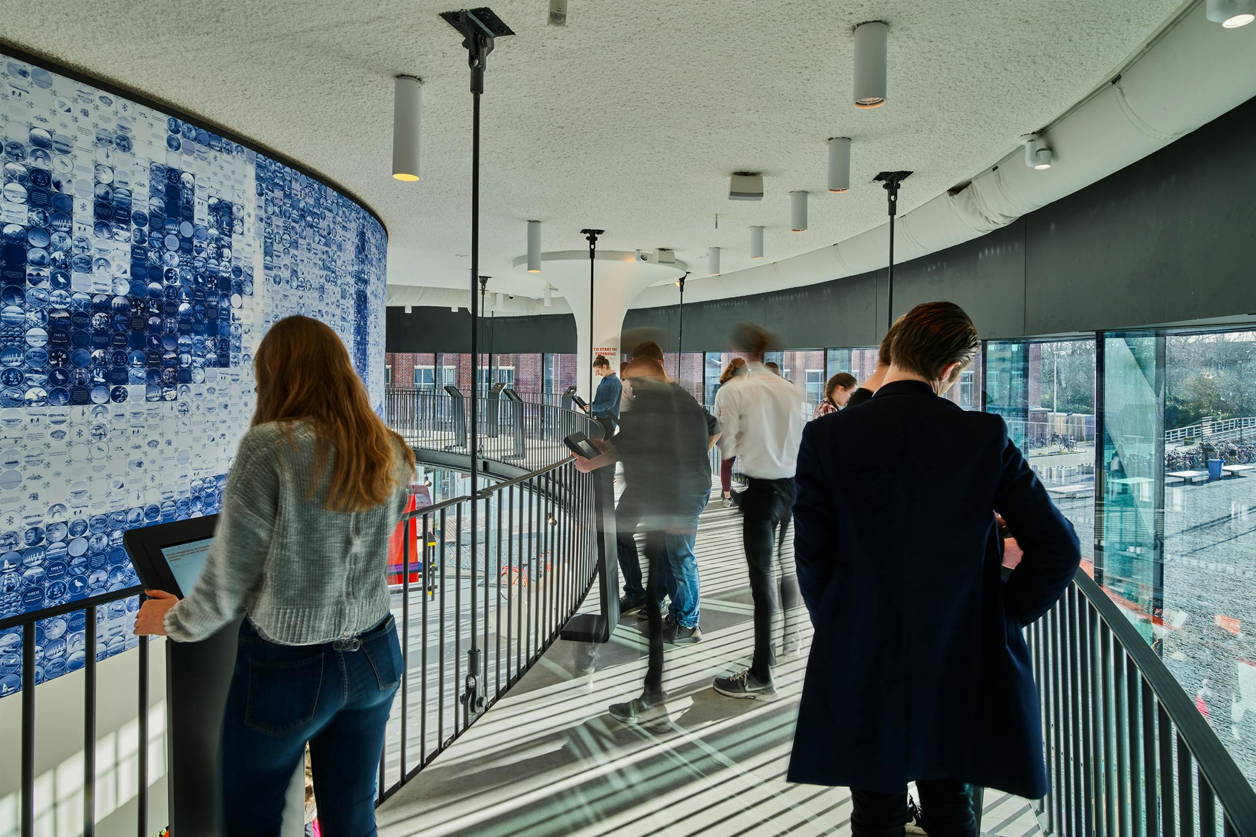 Οι άνθρωποι περπατούν και στέκονται σε ένα καμπύλο εσωτερικό μπαλκόνι με κιγκλίδωμα, που περιβάλλεται από μοντέρνα διακόσμηση και φωτιστικά.