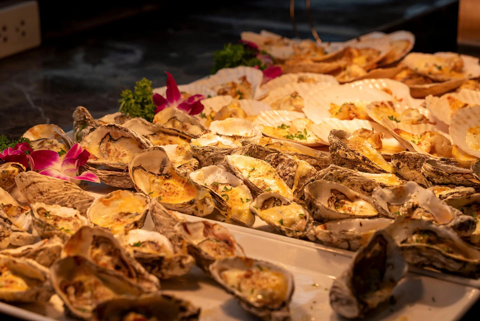 A close-up of a platter of baked oysters garnished with herbs and flowers, displayed on a serving table.