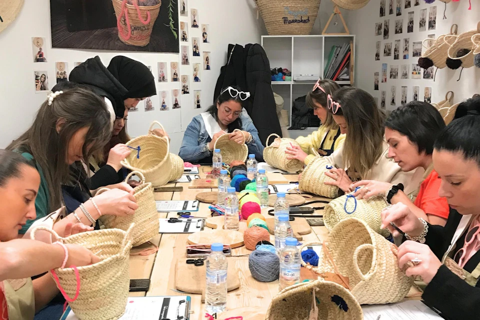 A group of women at a craft workshop, decorating straw baskets with colorful yarn and tools on a table.