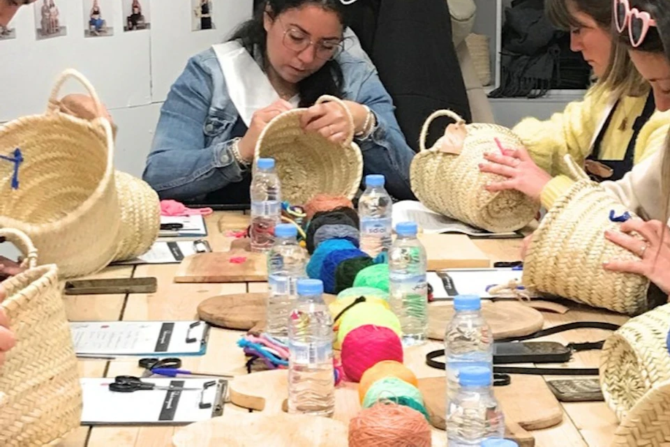 A group of women at a craft workshop, decorating straw baskets with colorful yarn and tools on a table.