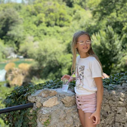 A young girl with glasses, holding a drink, stands by a stone wall with lush greenery in the background.