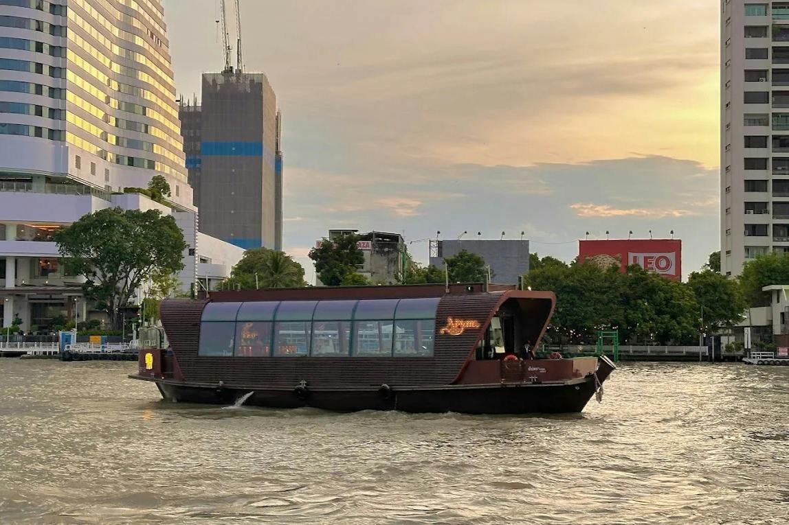 A modern riverboat with a wooden design and glass windows is moving on a city river during sunset.