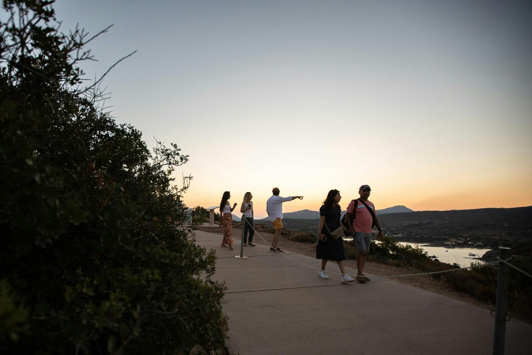 游客在苏尼奥角（ Cape Sounio ）朝日落方向行走。人们在褪色的天空下沿着低矮的灌木丛旁的小路漫步。