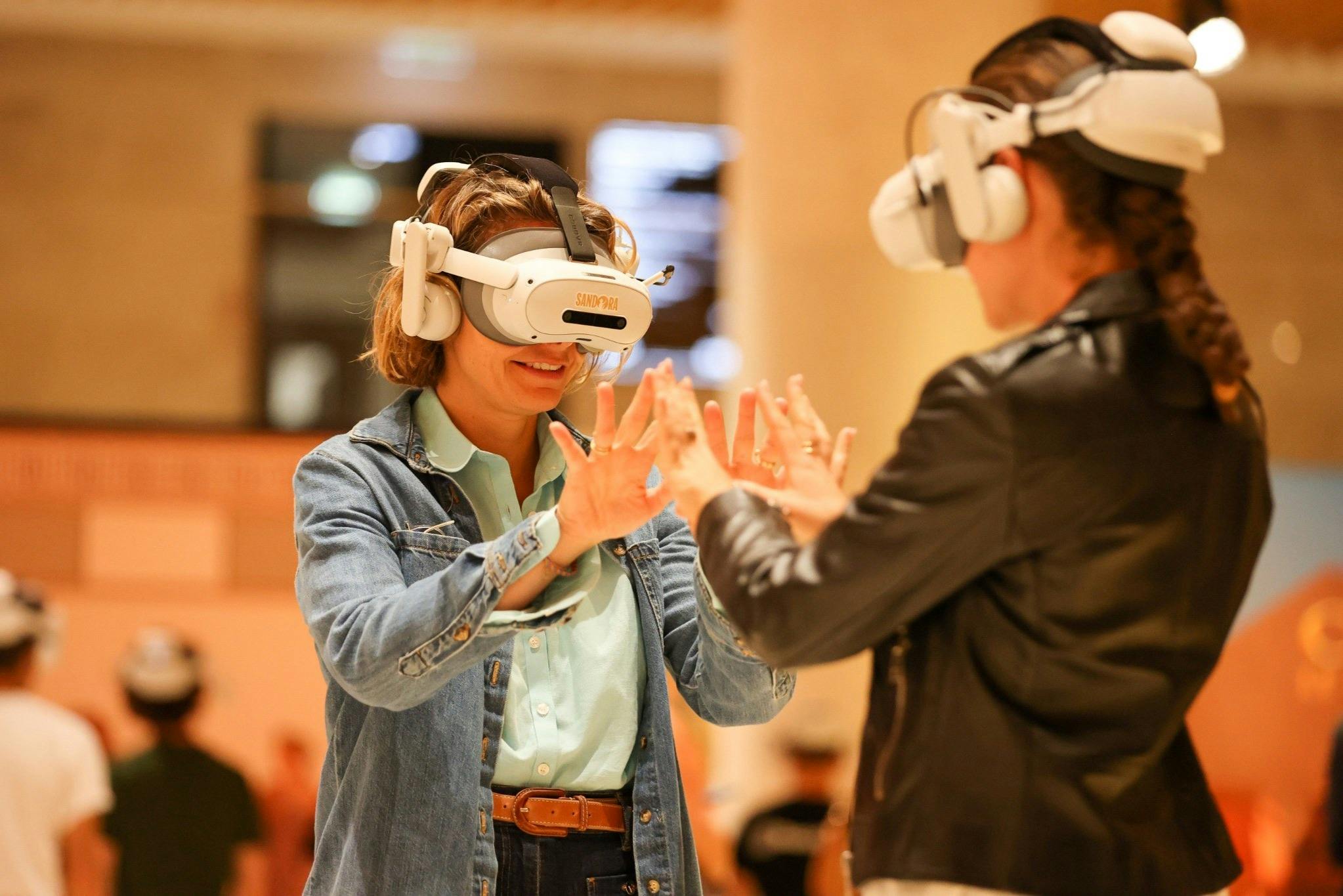 Two people wearing virtual reality headsets interact with each other in an indoor setting.
