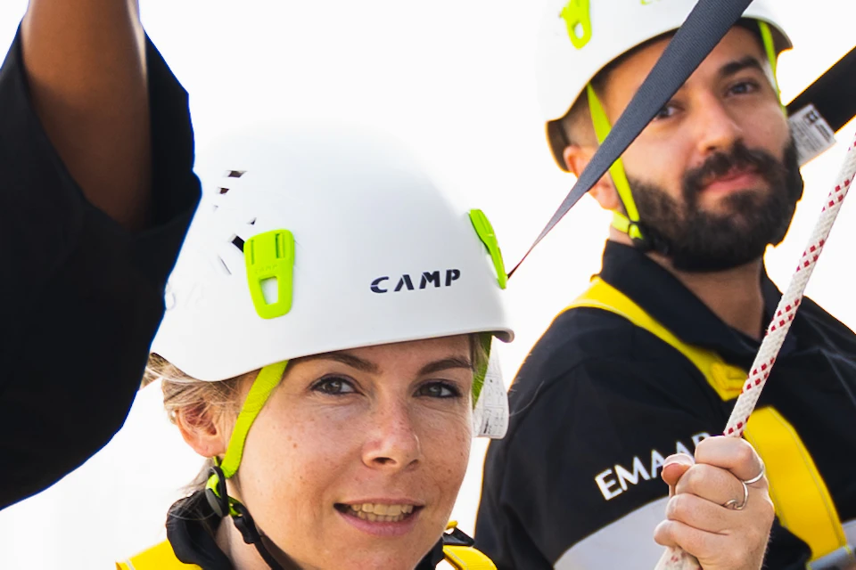 Five people in safety gear and helmets are attached to ropes on the edge of a high structure, smiling and looking at the camera.