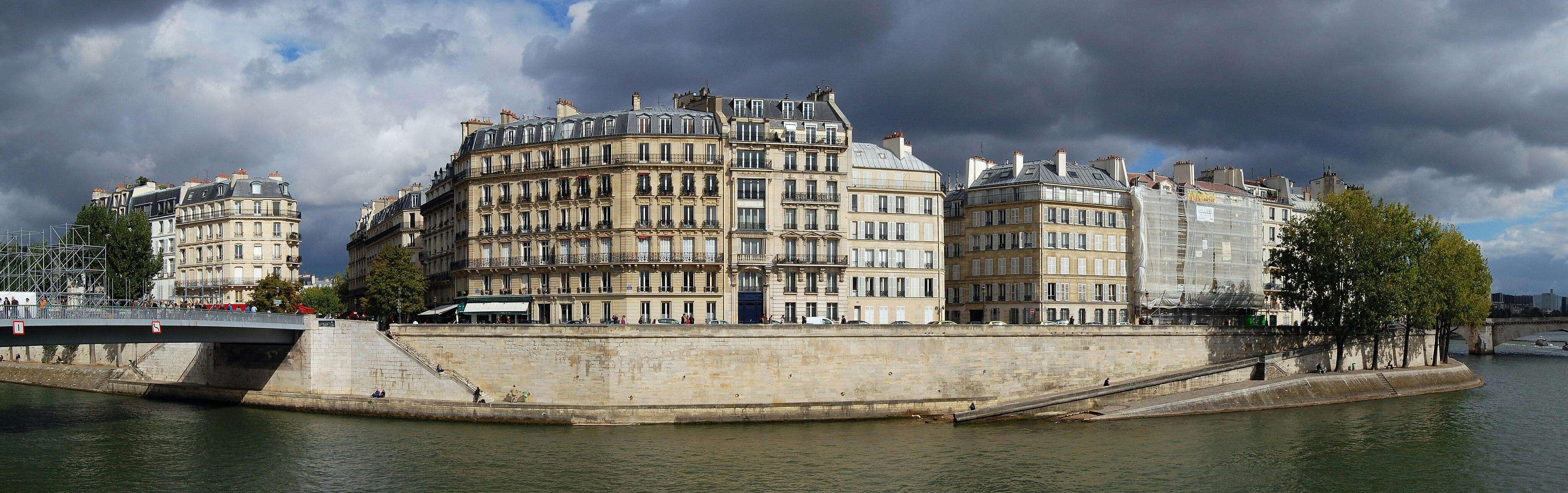 Row of Parisian buildings along the Seine River with a bridge and trees in the foreground under a cloudy sky.