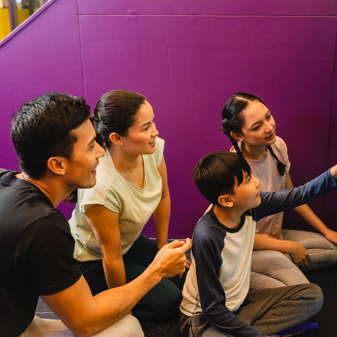 A group of four people, two adults and two children, sitting on the floor and smiling, interacting together indoors.