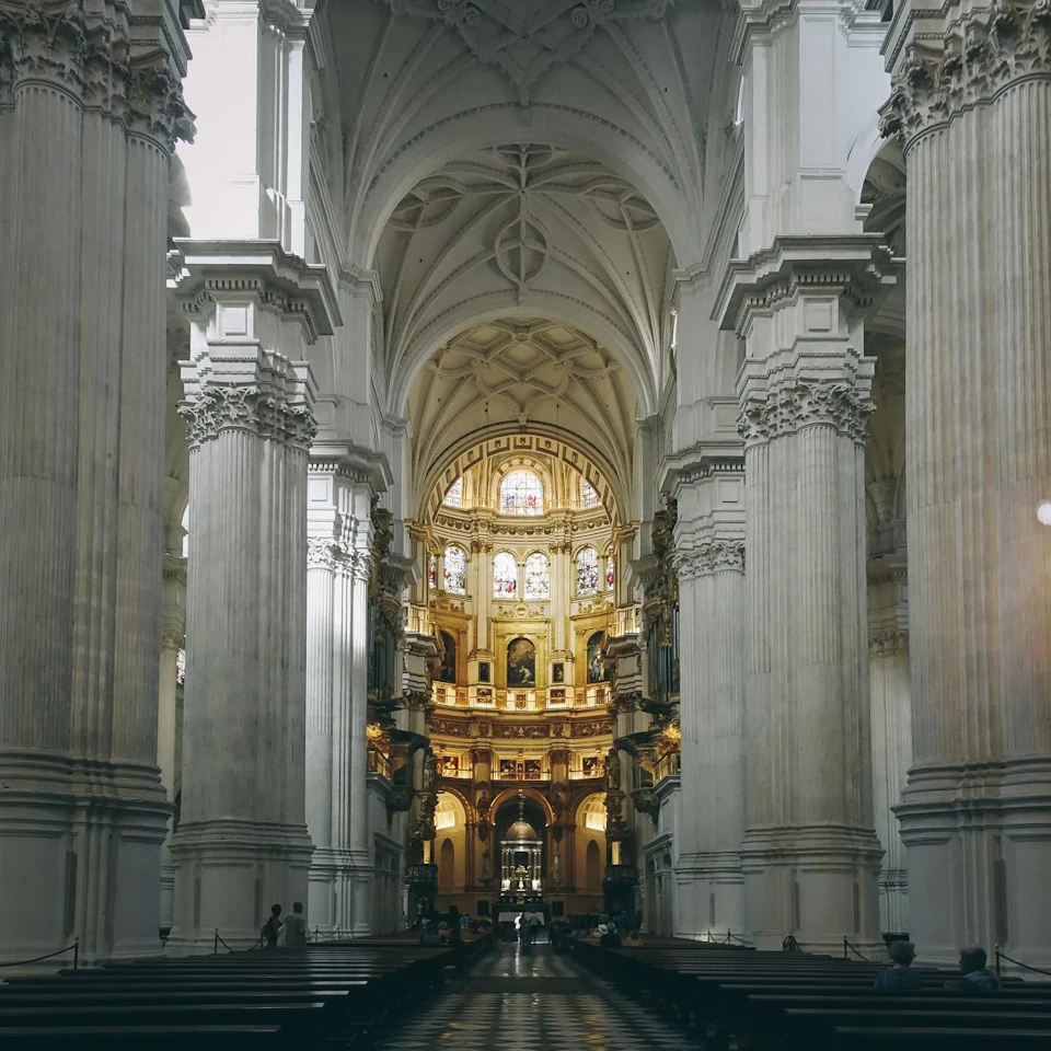 Ornate cathedral interior with tall arches, columns, and intricate stained glass windows above an illuminated altar.