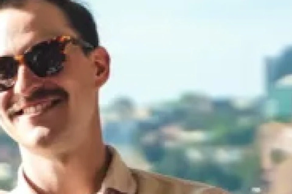 A man wearing sunglasses sits in a lounge area with drinks on the table, with a cityscape and bridge visible through the window.