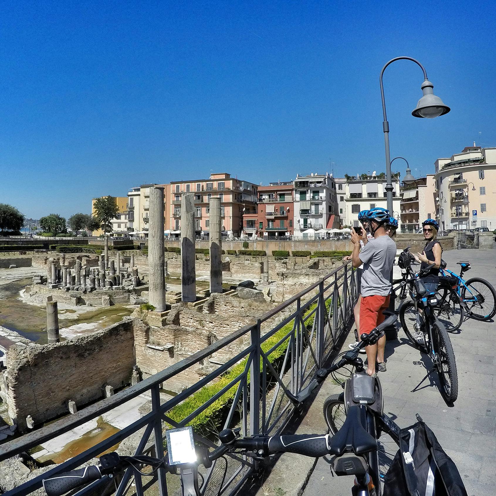 Ciclisti si fermano vicino ad antiche rovine con colonne, scattando foto. Gli edifici e un lampione sono visibili sotto un cielo azzurro e limpido.