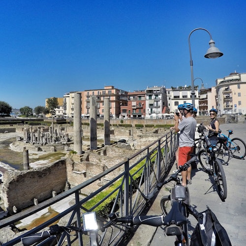 Cyclists stop near ancient ruins with columns, taking photos. Buildings and a streetlamp are visible under a clear blue sky.