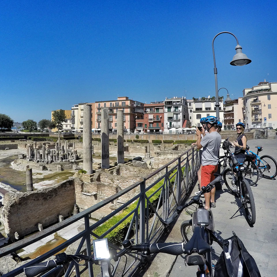Cyclists stop near ancient ruins with columns, taking photos. Buildings and a streetlamp are visible under a clear blue sky.