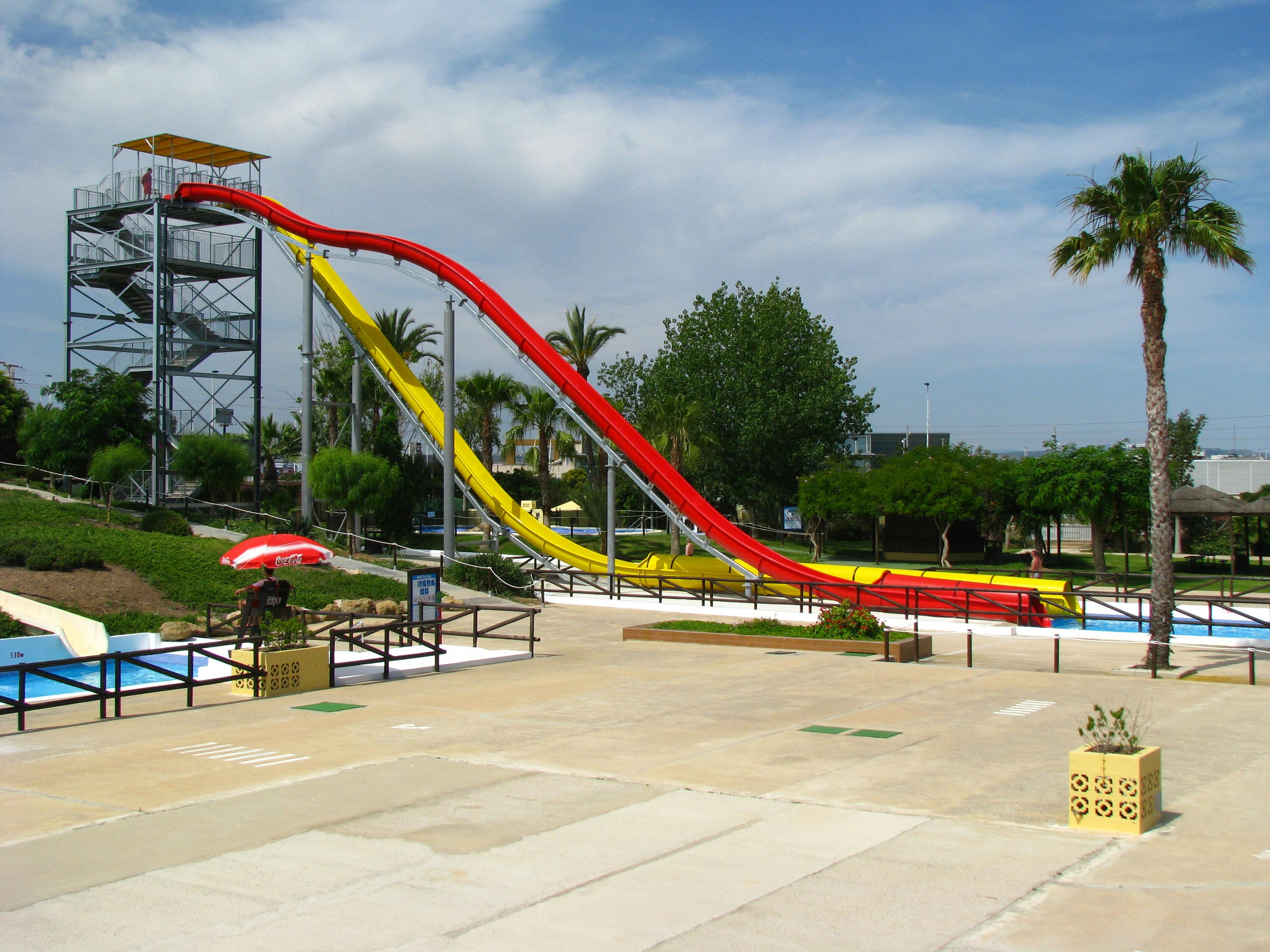 Un alto scivolo con sezioni rosse e gialle, circondato da alberi e da un cielo azzurro in un parco acquatico.
