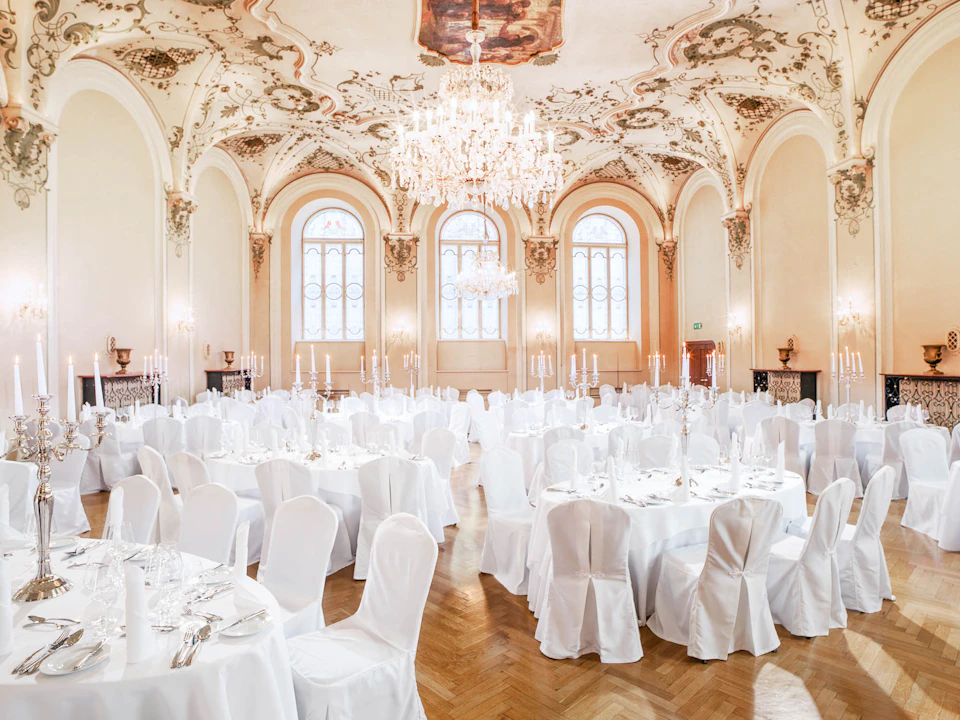 An elegant ballroom set for an event, with round tables covered in white cloths, white chair covers, chandeliers, and ornate ceiling designs.