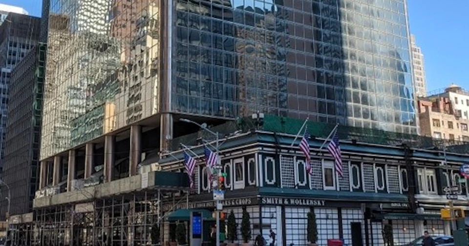 A city street corner with a restaurant building decorated with U.S. flags, set against a backdrop of glass skyscrapers.