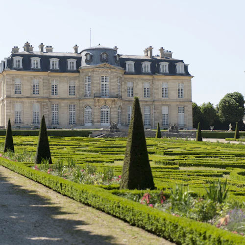 En storslagen historisk byggnad med välskötta trädgårdar med trimmade häckar och koniska buskar i förgrunden.