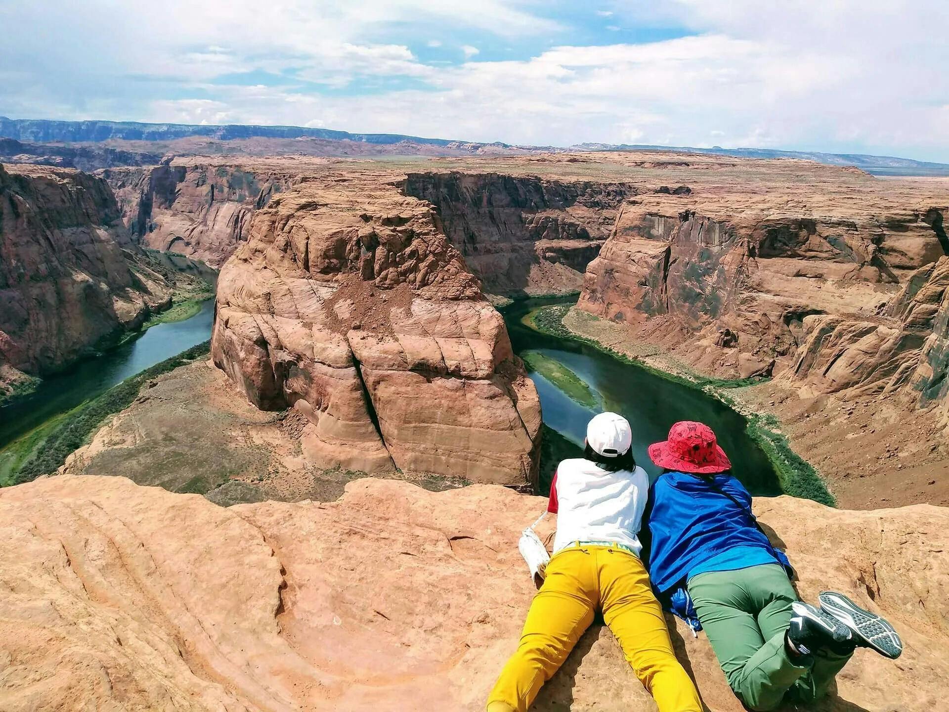 Two people in colorful clothes lie on the edge of a canyon, overlooking a river winding through the rocky landscape.