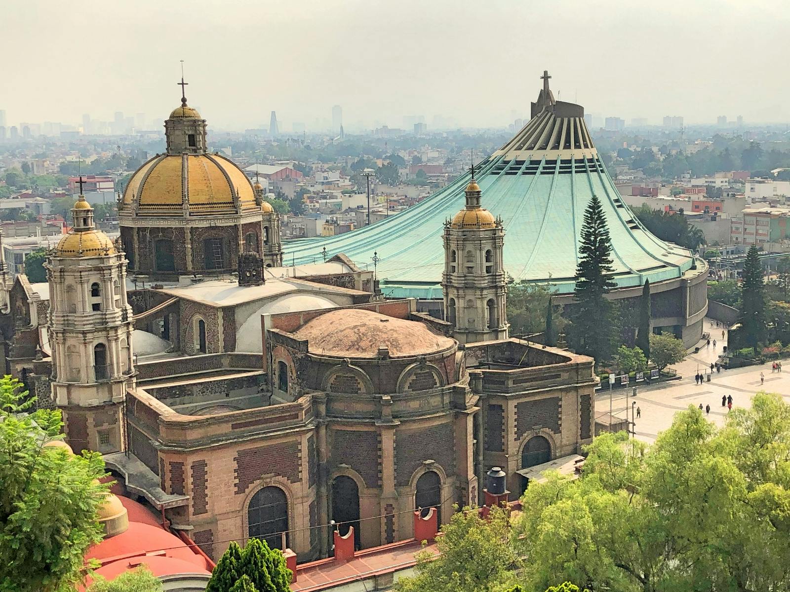 Historický kostel s kupolí a moderní bazilikou se šikmou střechou v městském pozadí. Stromy v popředí, panorama vybledlé oparem.