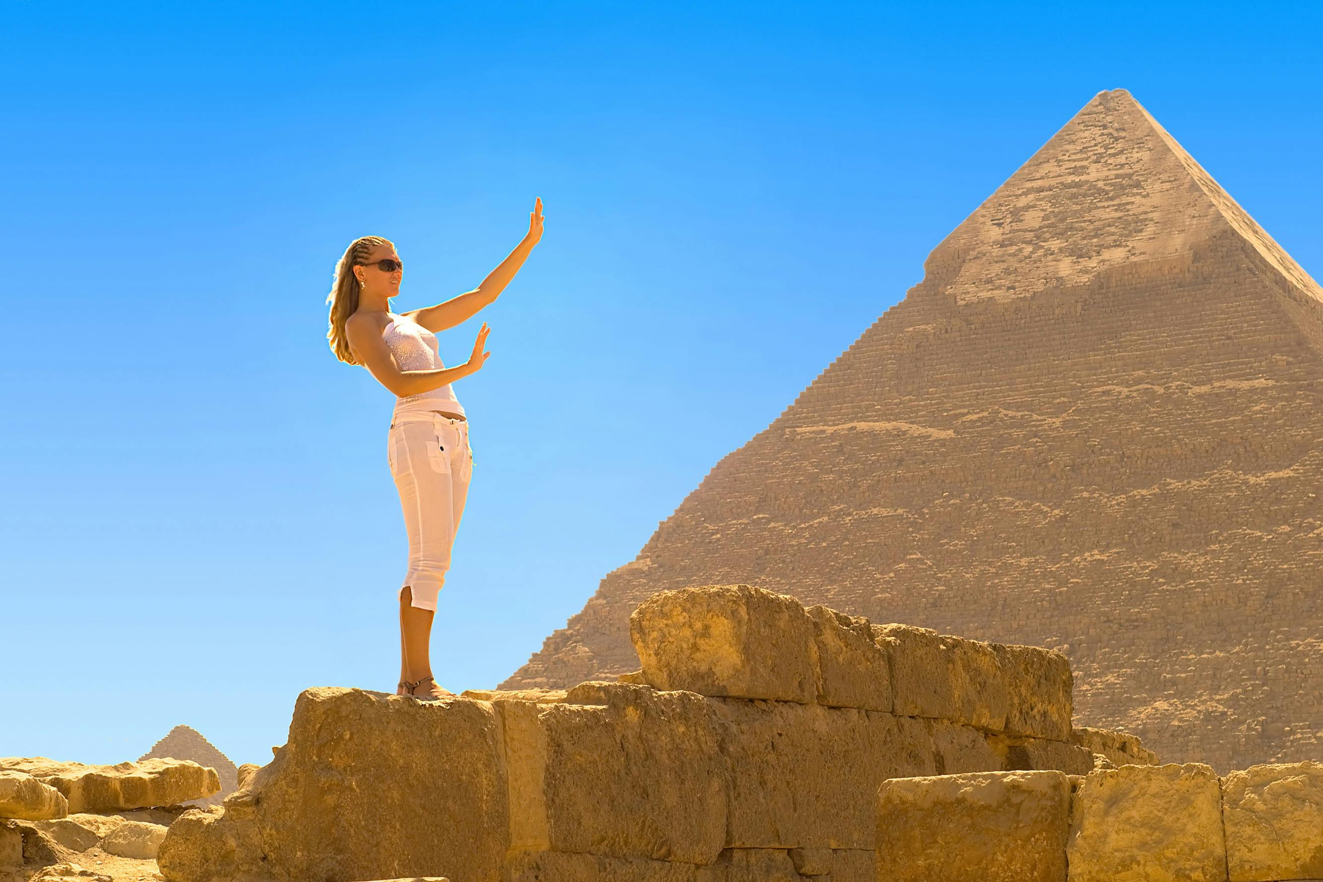 A woman in white clothing stands on a stone structure with the pyramids in the background under a clear blue sky.