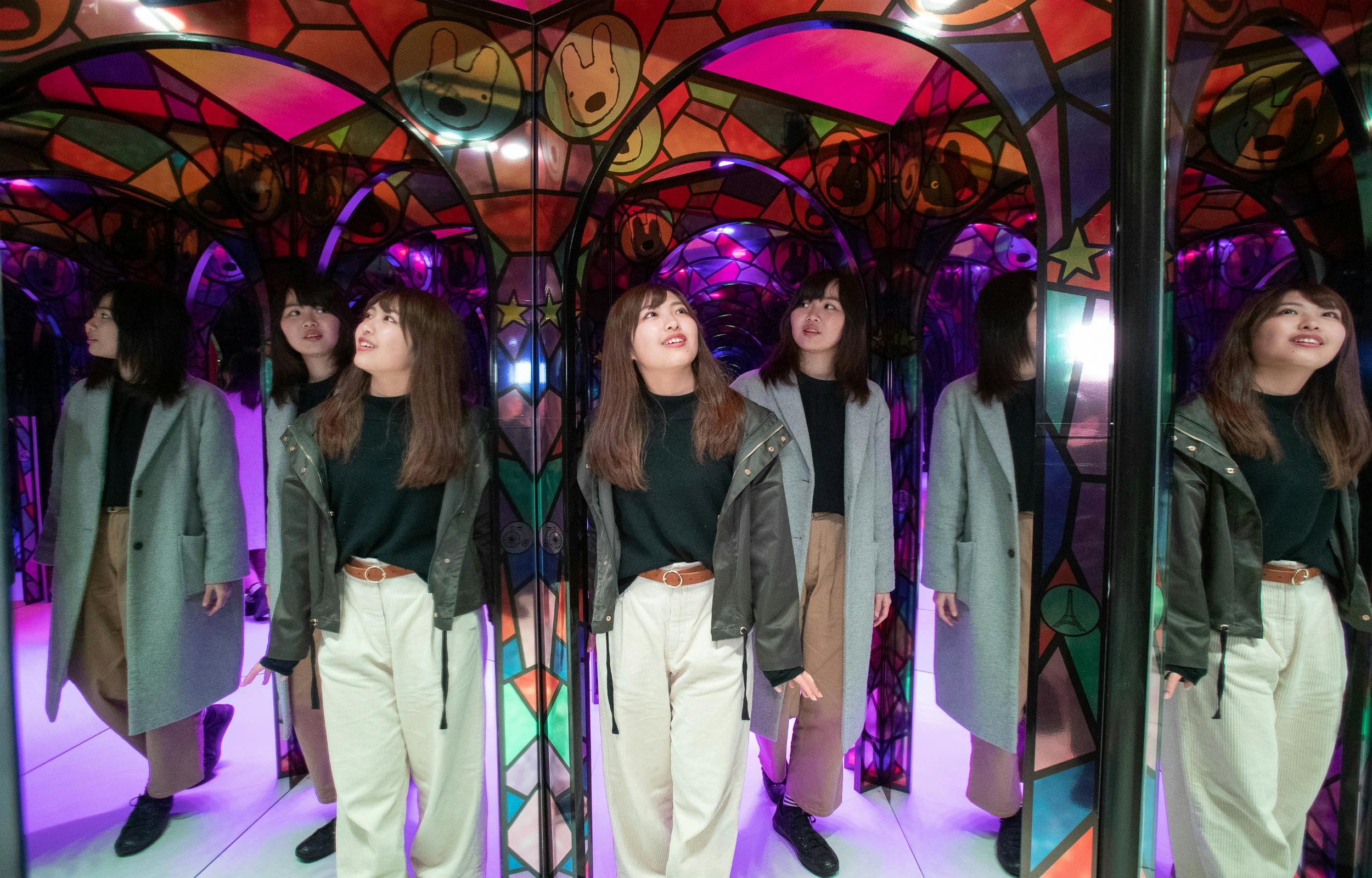 一位女士站在五彩缤纷的镜像隧道中，产生了多重反射。 她穿着浅色裤子、深色上衣和外套。