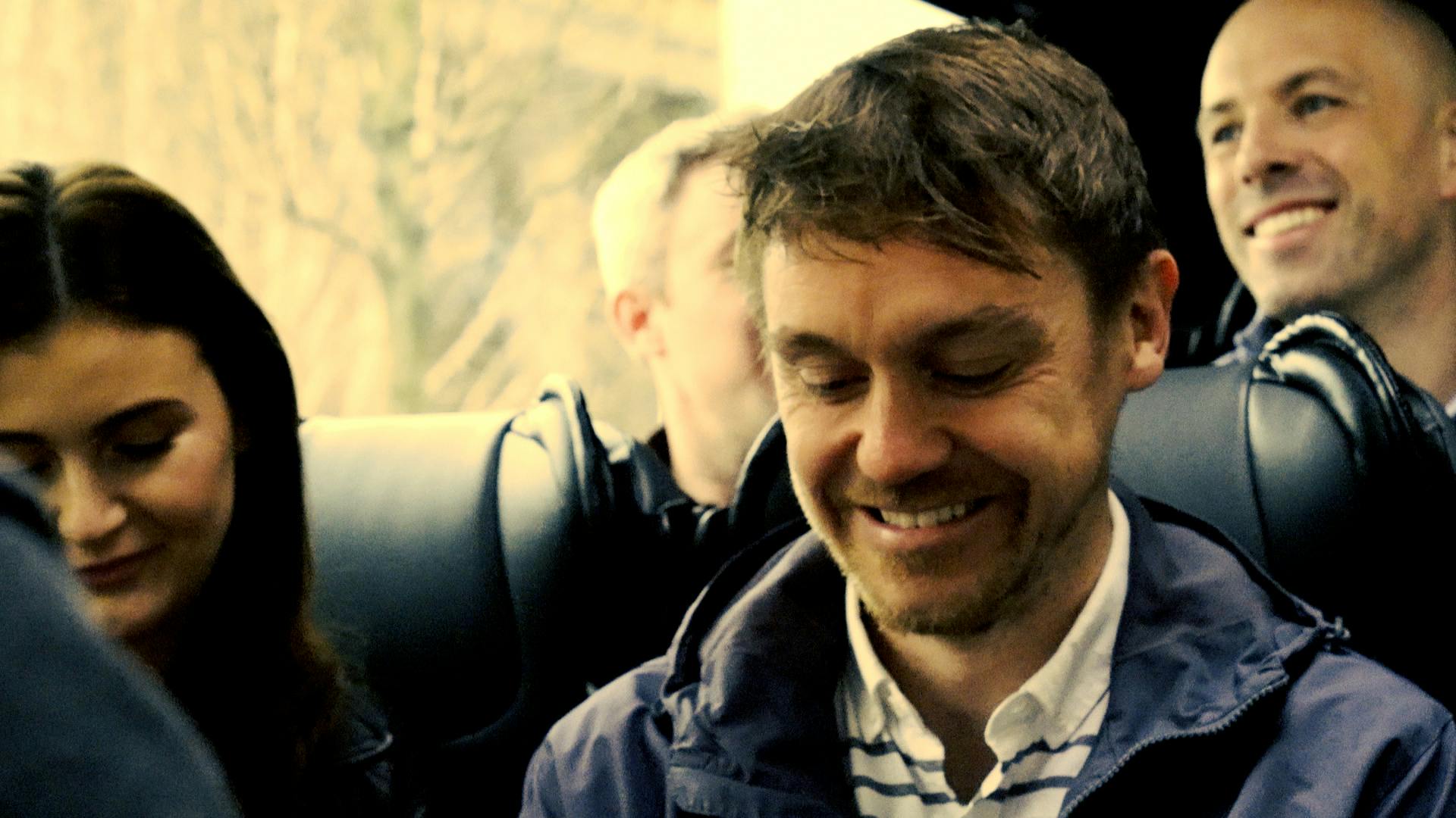 一个棕色短发，蓄着微微胡须的男人低头微笑，背景中隐约可见另一个人。