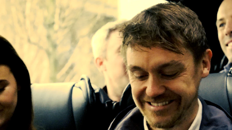 A man with short brown hair and a slight beard smiles while looking down, with another person faintly visible in the background.