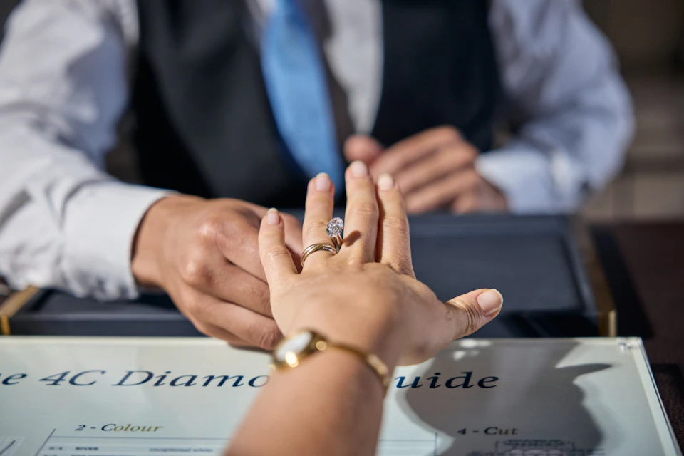 A person wearing a ring is having their hand examined by another person, with a document labeled 'Diamond Guide' visible.