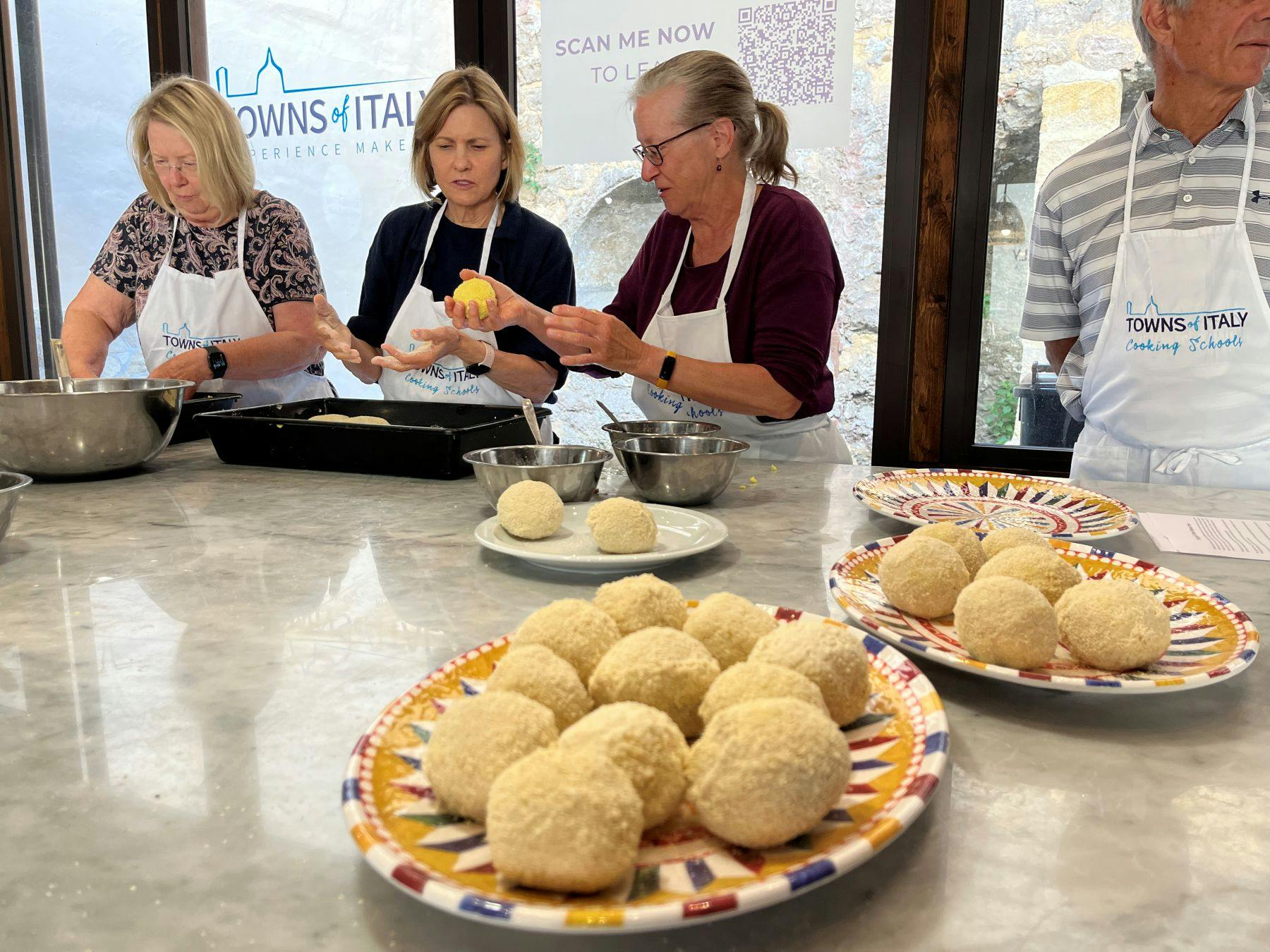 Three women wearing aprons are preparing food around a counter with bowls, plates, and dough balls on it.
