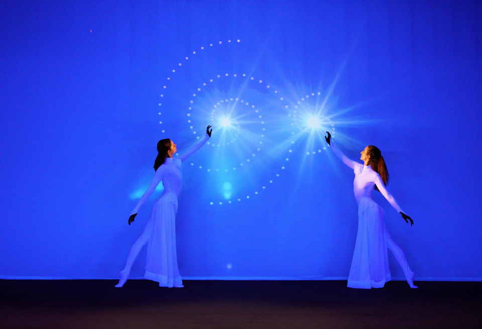 Two performers in white costumes reach out towards glowing, spiral light patterns on a blue background.