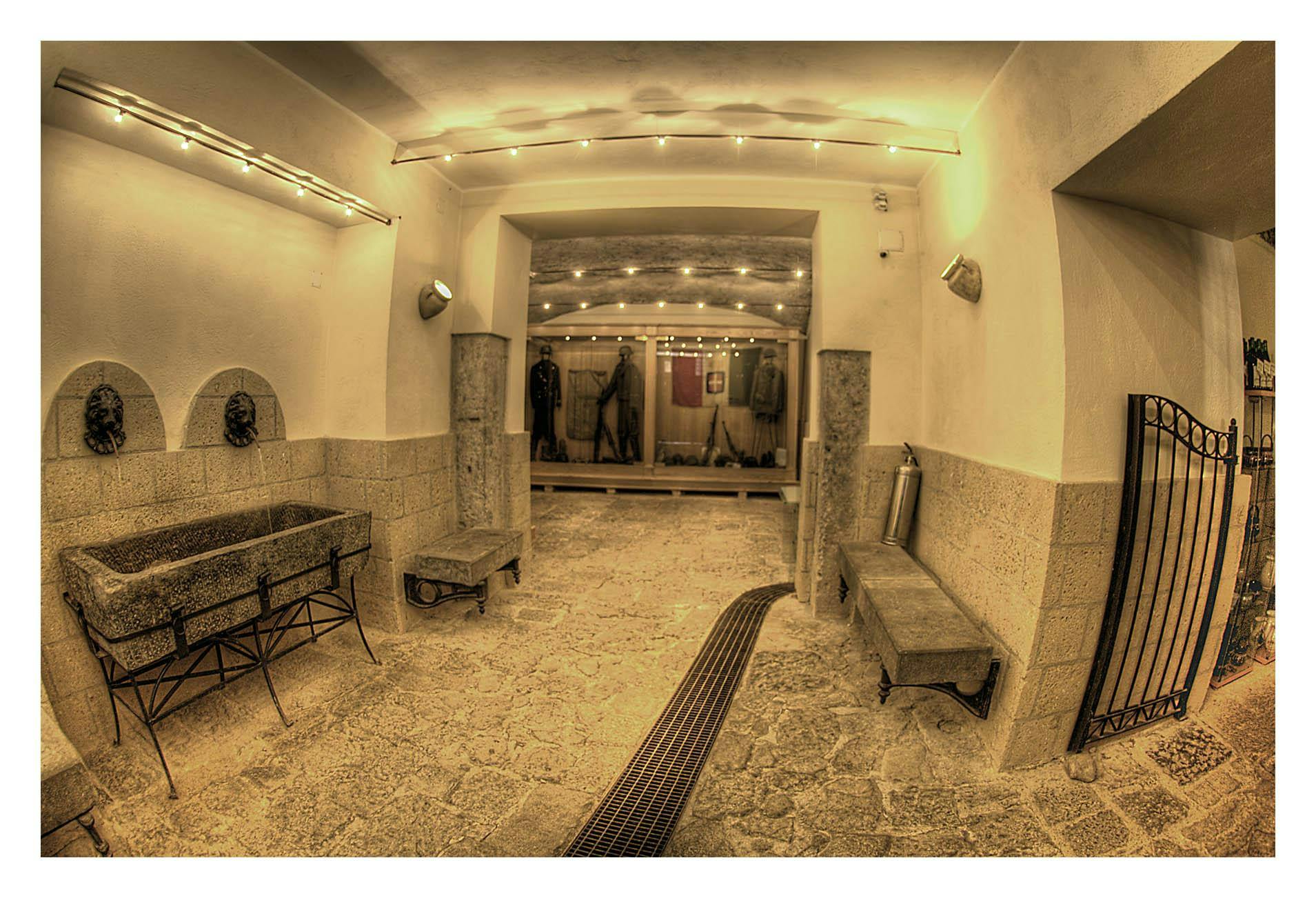 Dobře osvětlená kamenná chodba s lavičkami, vodními fontánami ve tvaru lva a figurínami v uniformách vystavenými za sklem.