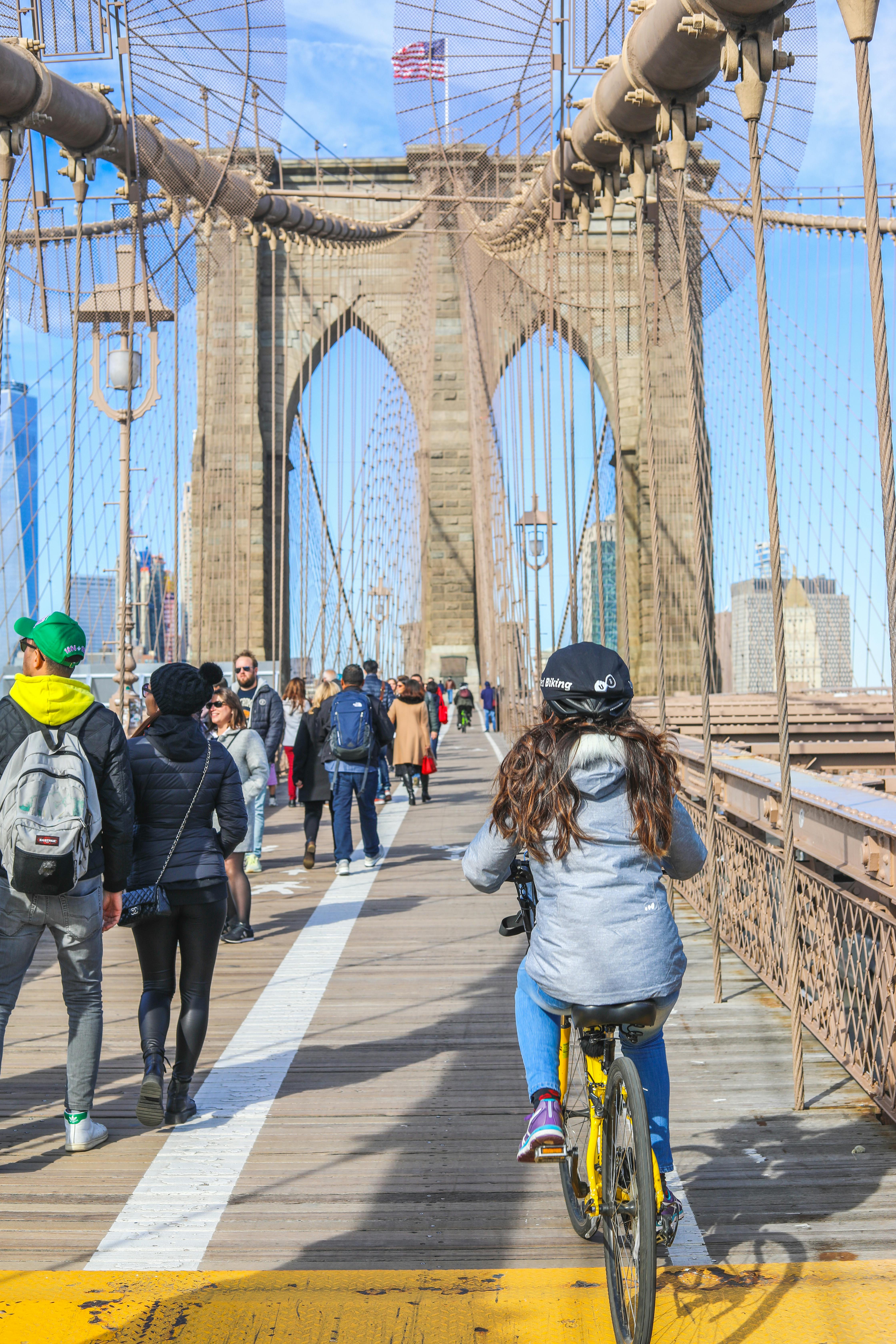 Persone che camminano e vanno in bicicletta su un ponte con grandi archi di pietra e cavi sotto un cielo azzurro e limpido.
