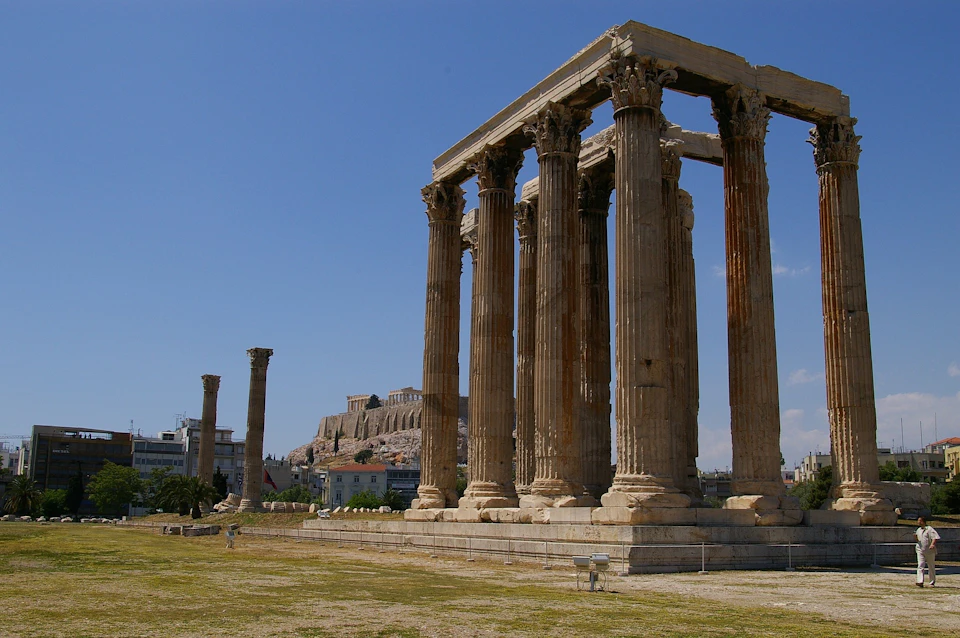 Ancient Greek columns with missing sections stand in an open area, with Acropolis of Athens visible in the background. Clear sky above.
