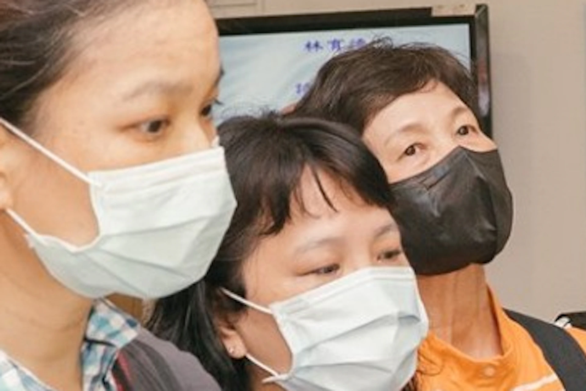 A group of people wearing face masks standing indoors, some listening attentively, in a brightly lit room.