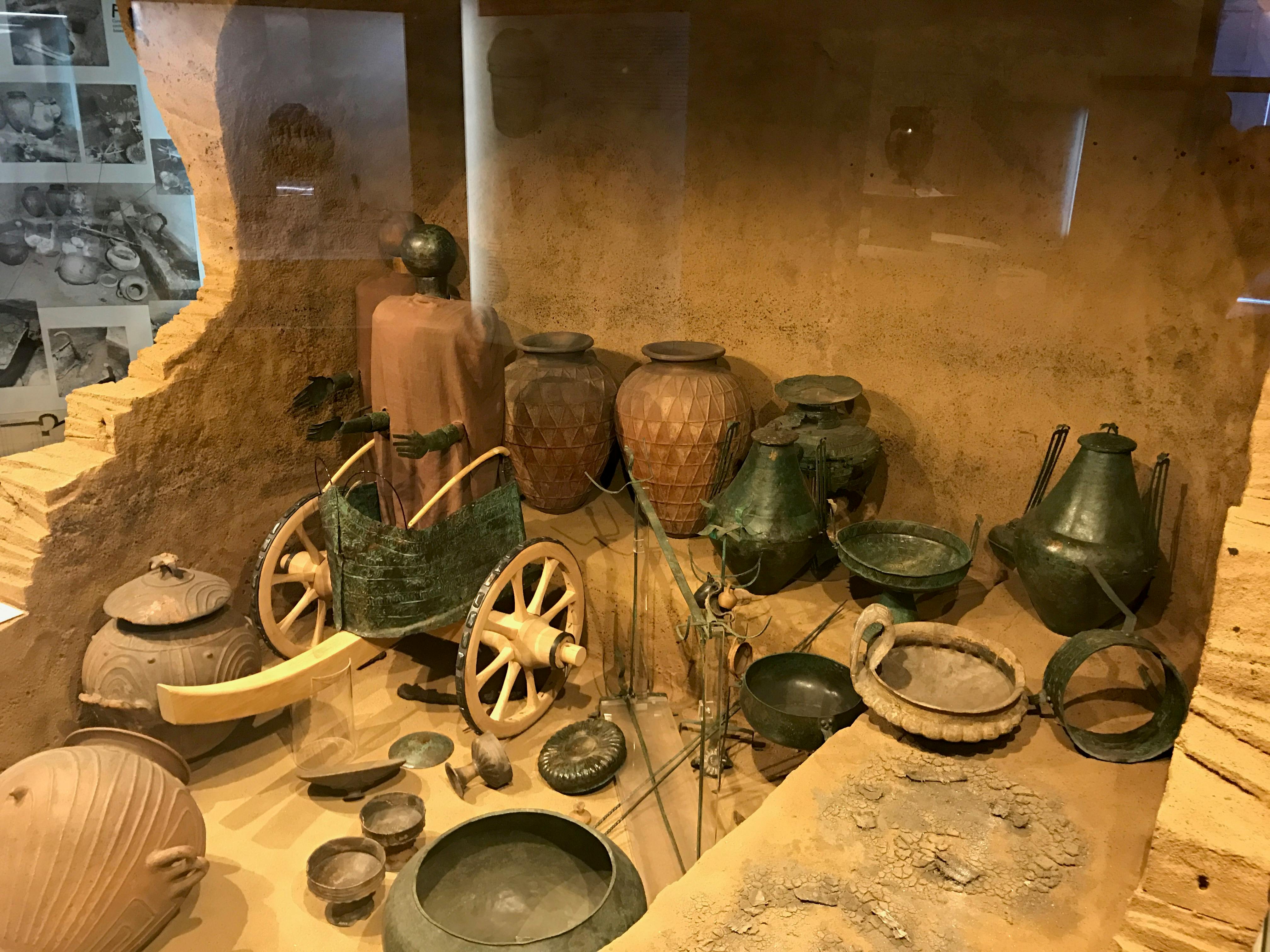 Een museumvitrine met oud aardewerk, metaalwerk, een miniatuur strijdwagen en verschillende artefacten tegen een zanderige achtergrond.