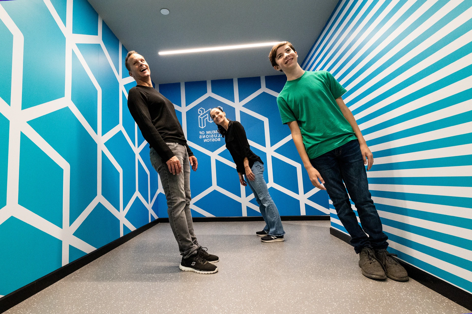 Three people lean at an angle in a blue room with geometric patterns at the Museum of Illusions in Boston.