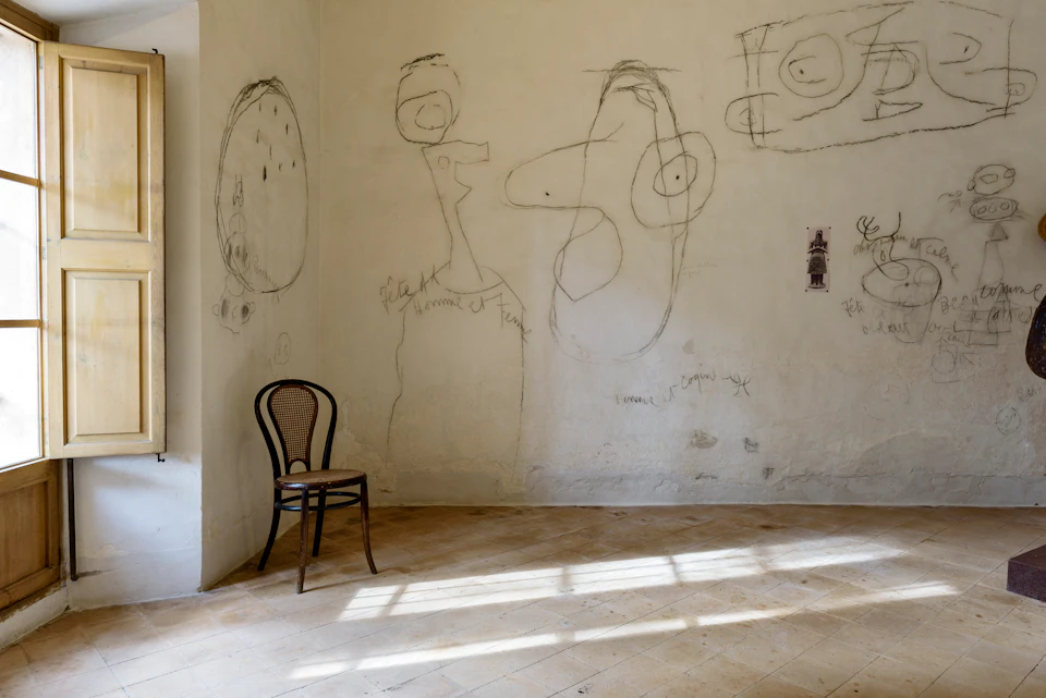 A single wooden chair sits in a well-lit room with simple, abstract drawings on its white walls.