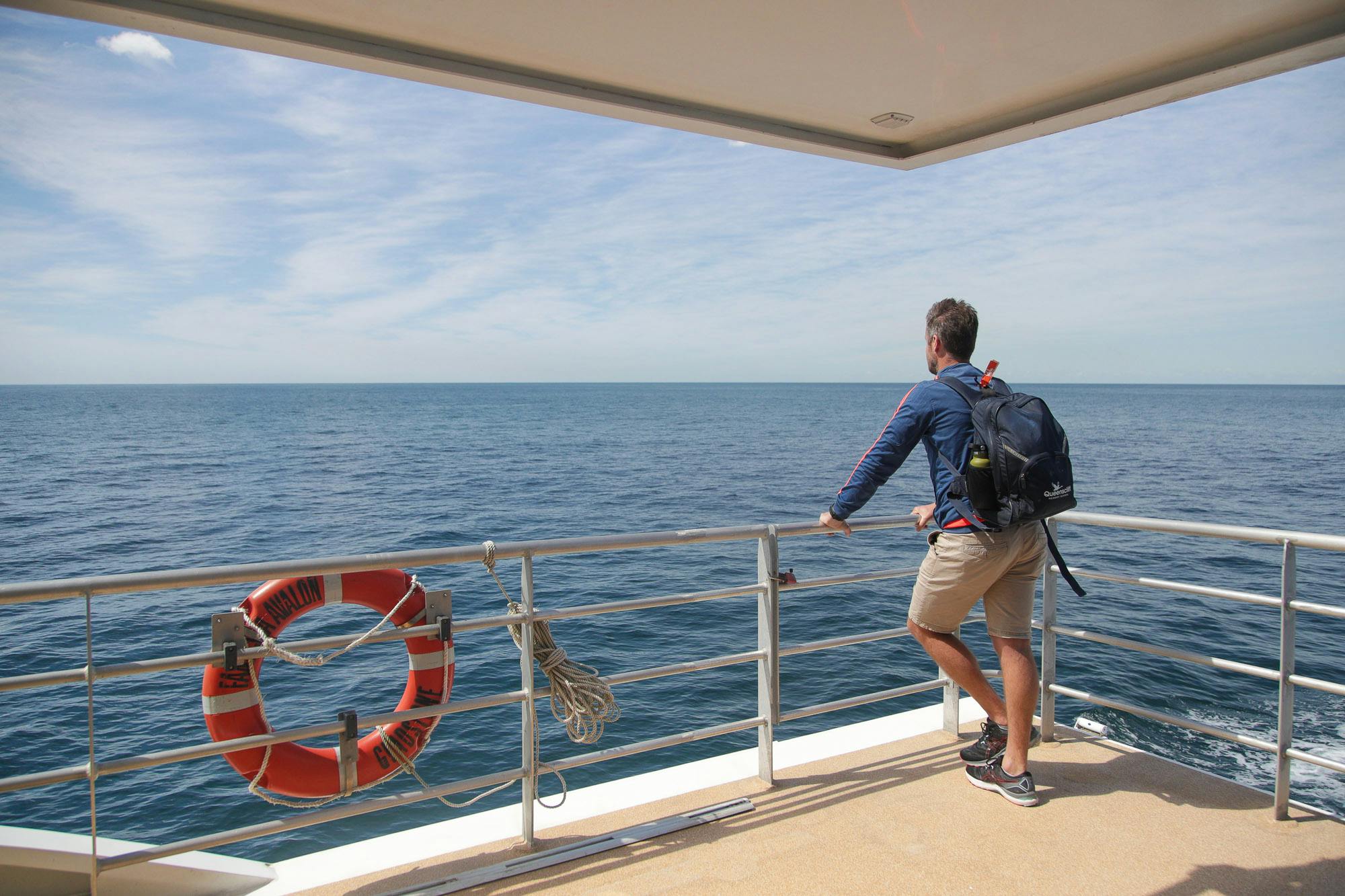 A person with a backpack stands on a boat deck, leaning on a railing and looking out at the ocean under a partly cloudy sky.
