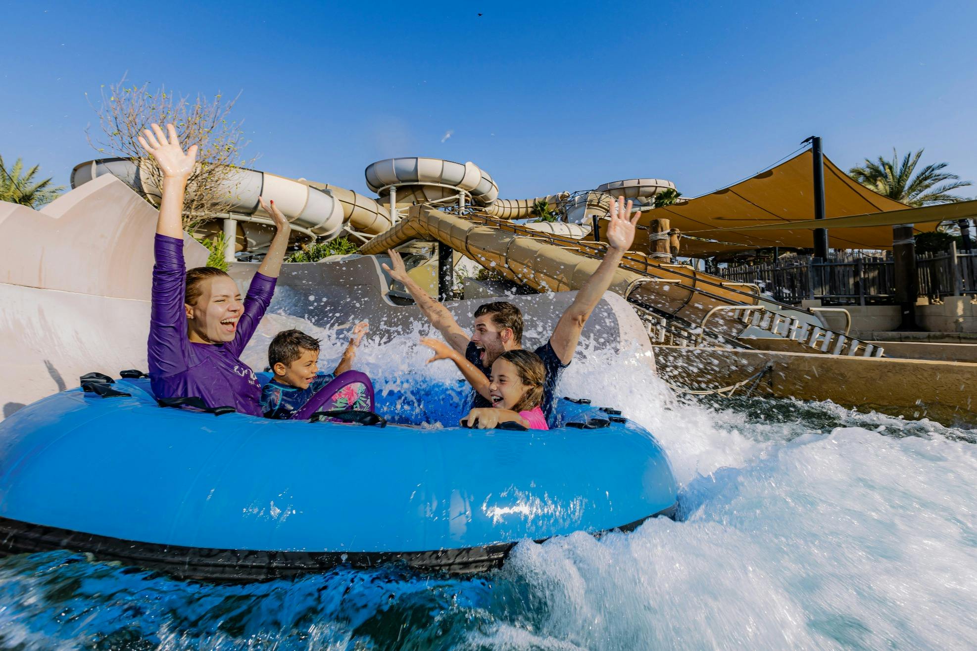Une famille de quatre personnes profite d'un toboggan aquatique sur un radeau gonflable bleu sous un soleil radieux, les bras levés en signe d'excitation.