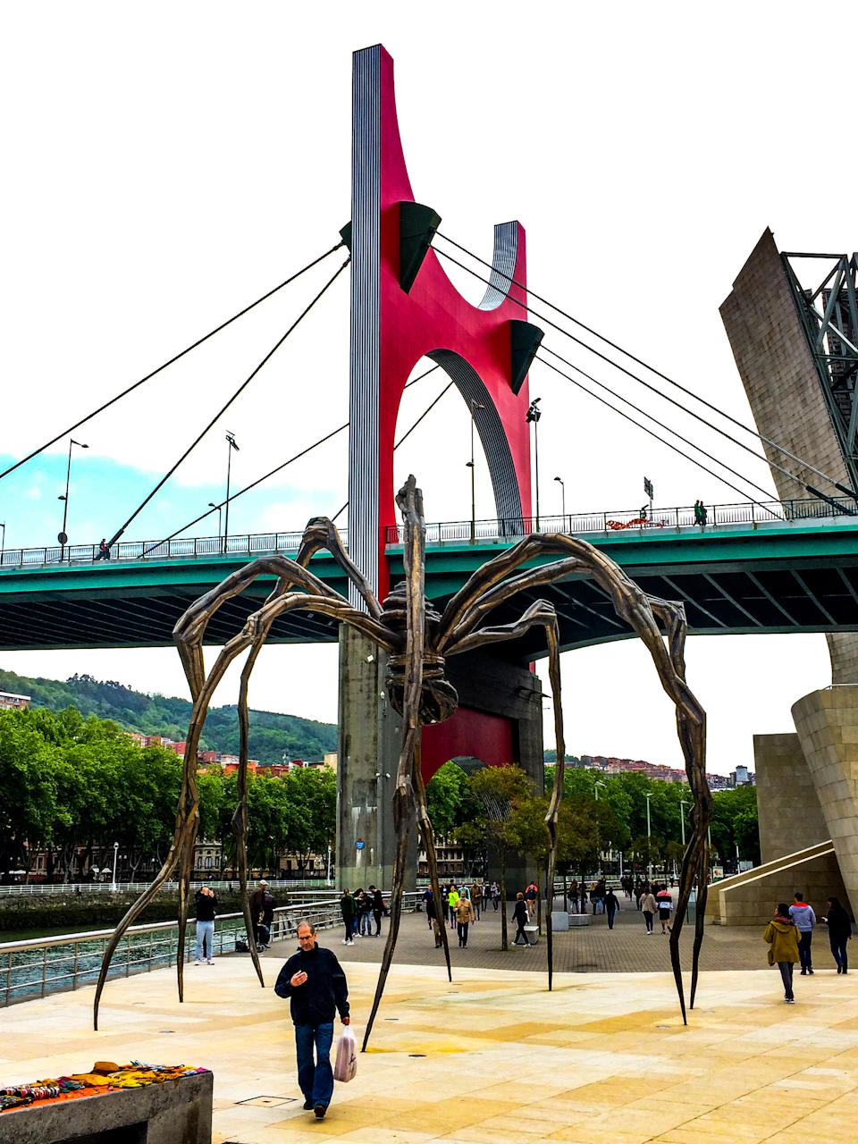 A large spider sculpture near a bridge with a red arch, surrounded by people in an urban park with trees and buildings in the background.