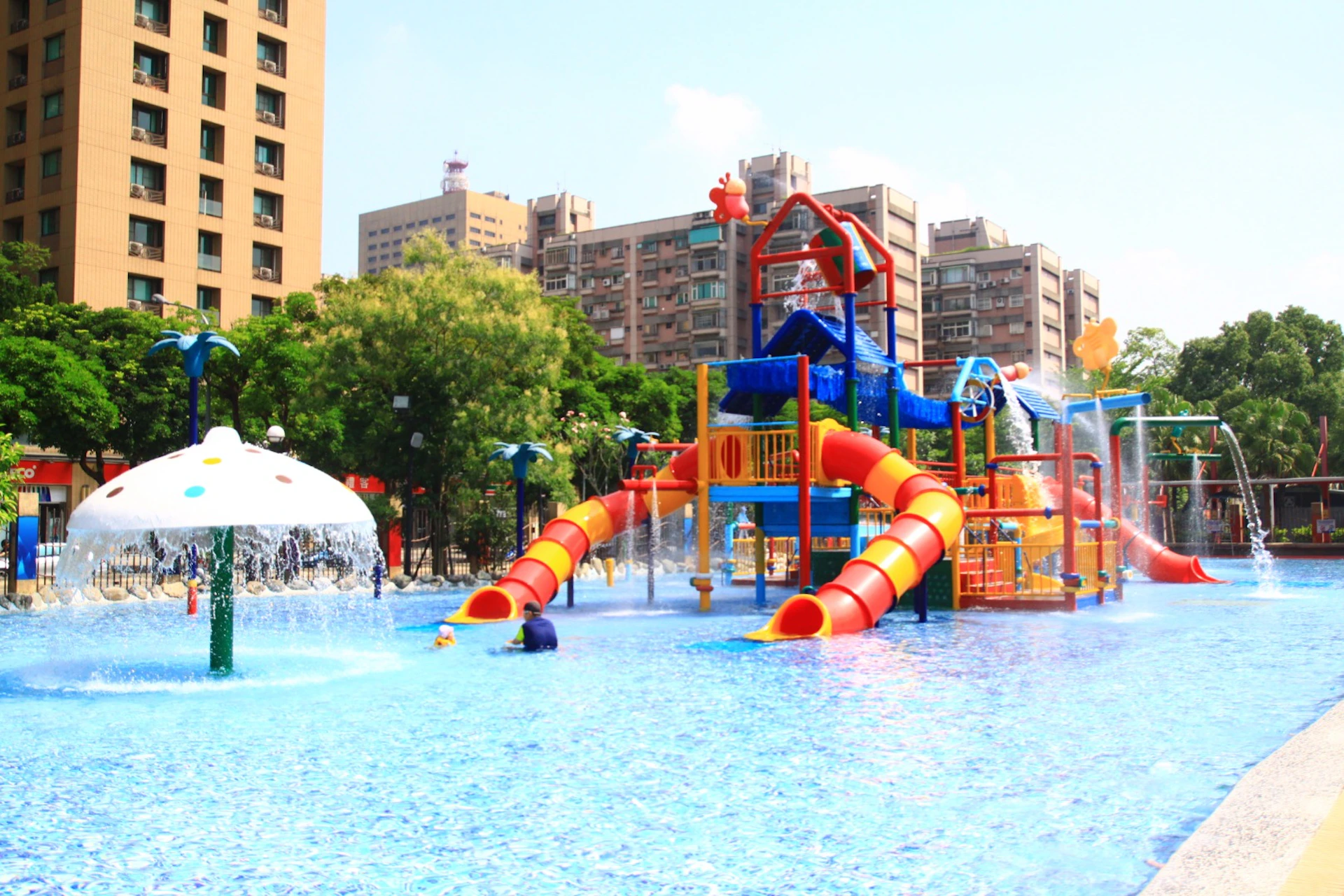 A colorful water playground with slides and structures in a pool, surrounded by trees and tall buildings under a clear sky.