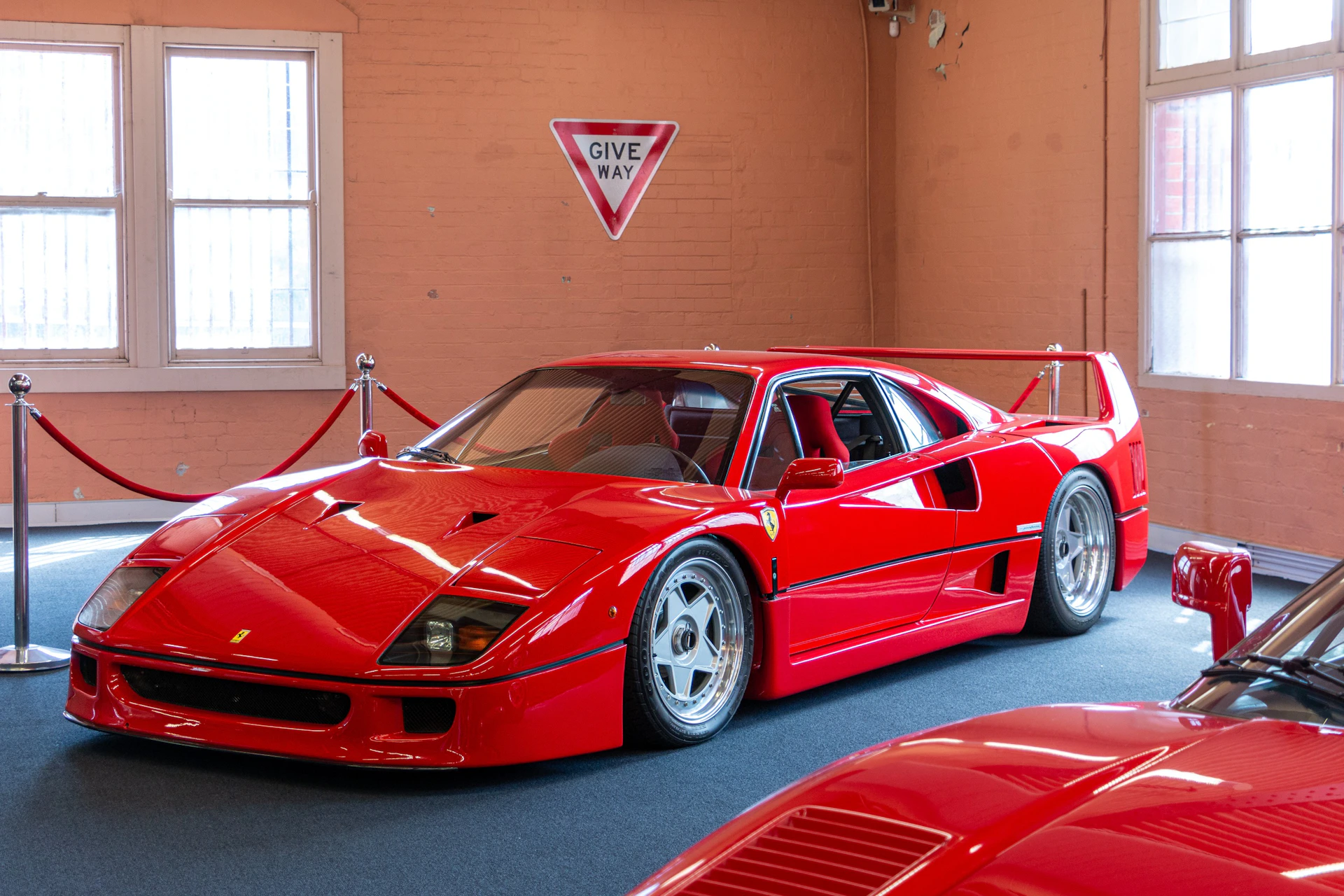 A red sports car displayed indoors, with a 'Give Way' sign on an orange wall behind it.