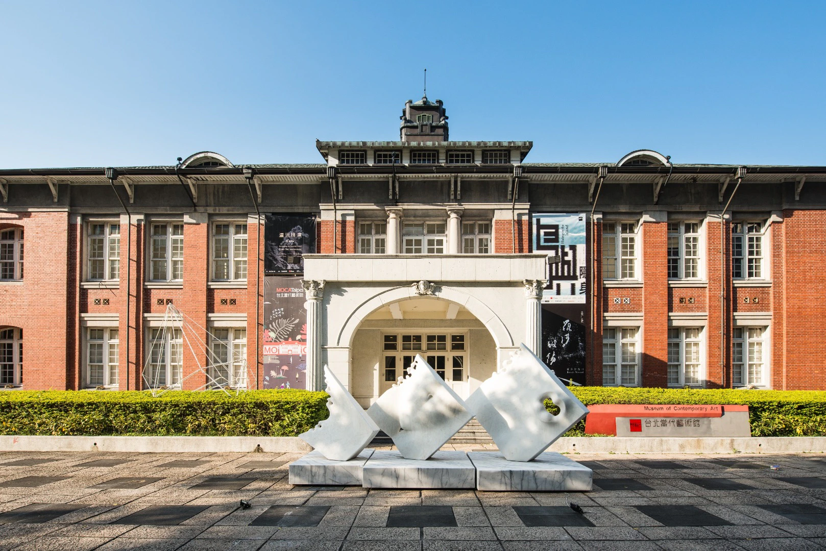 A historic brick building with white columns and a modern sculpture in the foreground under a clear blue sky.