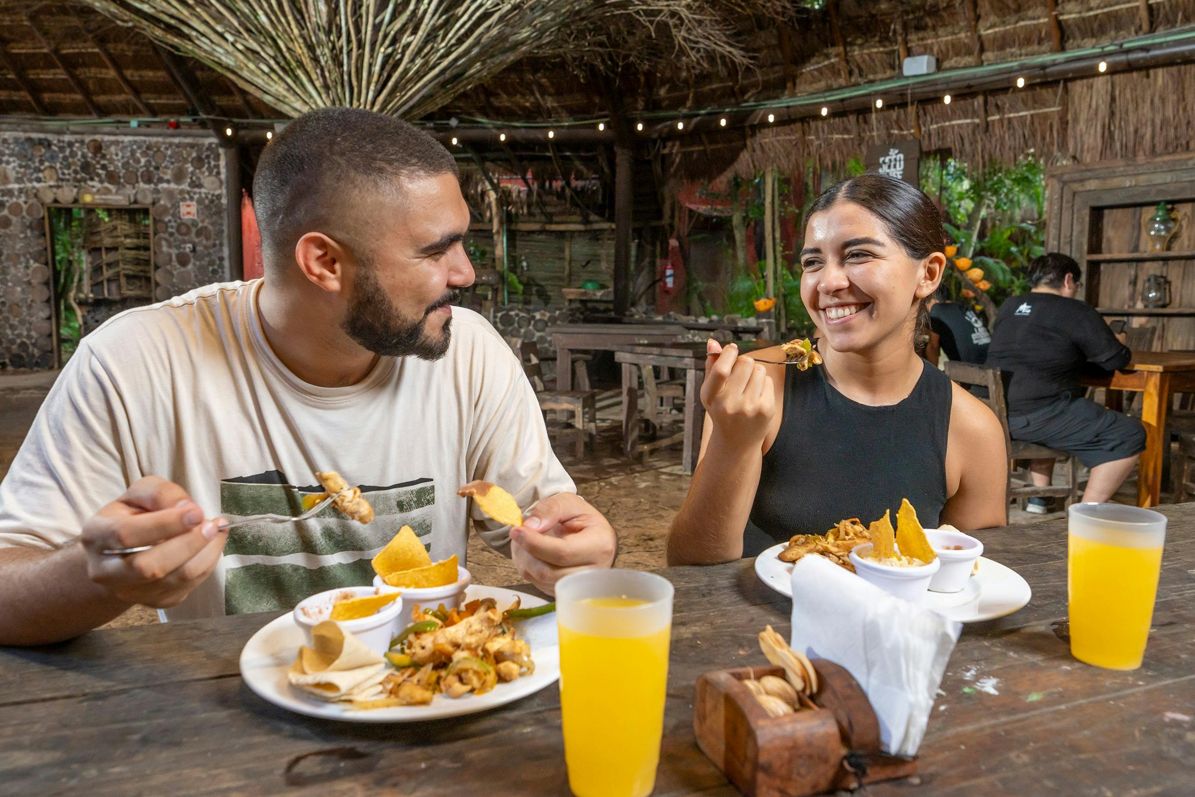 木製の家具とストリングライトを備えた素朴な野外レストランで、2人が一緒に笑顔で食事をしています。